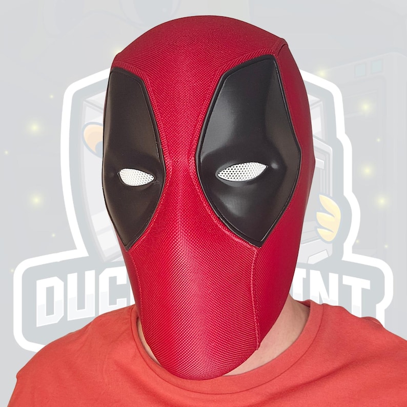 May include: A red Deadpool mask with black eye coverings and white mesh eye openings. The mask is made of a textured material and covers the entire head. The person wearing the mask is wearing a red shirt.