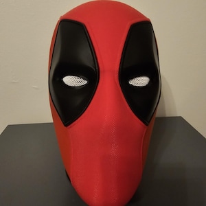 May include: A red and black Deadpool mask. The mask features black eye coverings with white mesh eye openings. The rest of the mask is red. The mask is sitting on a dark gray surface.