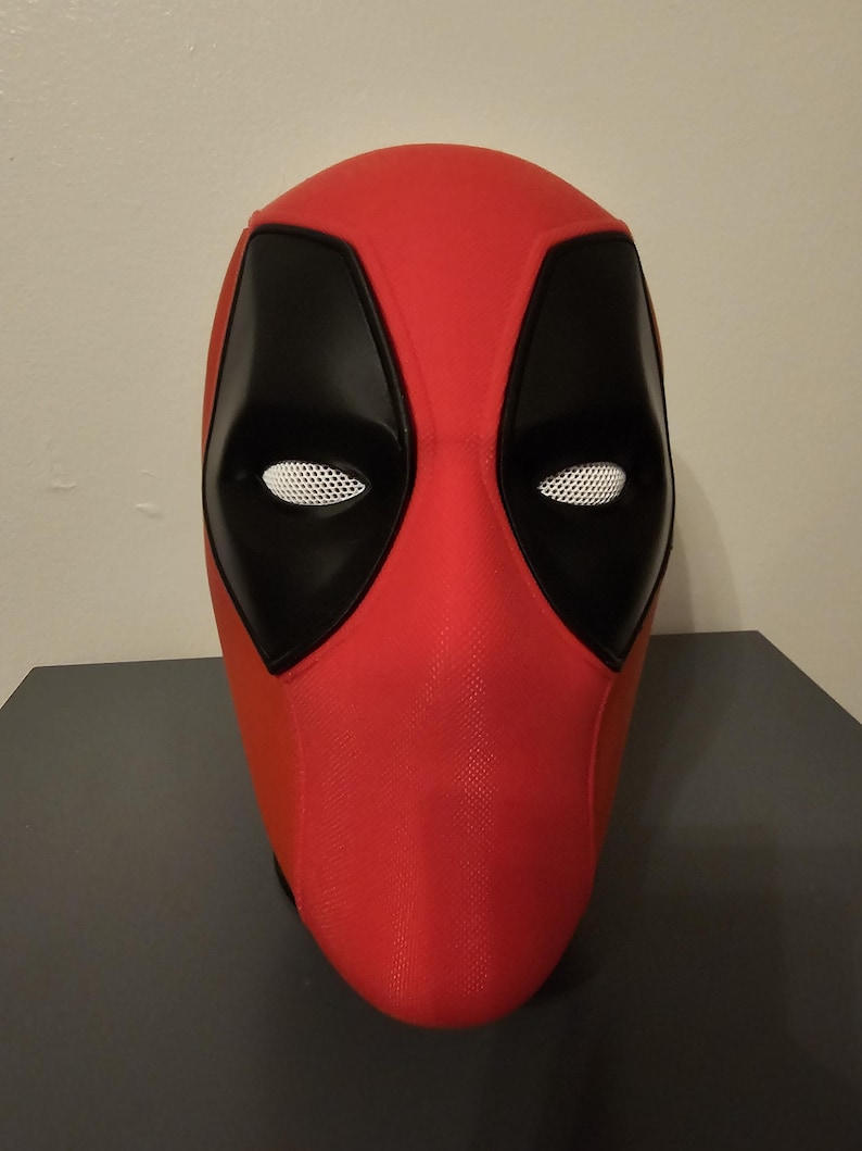 May include: A red and black Deadpool mask. The mask features black eye coverings with white mesh eye openings. The rest of the mask is red. The mask is sitting on a dark gray surface.
