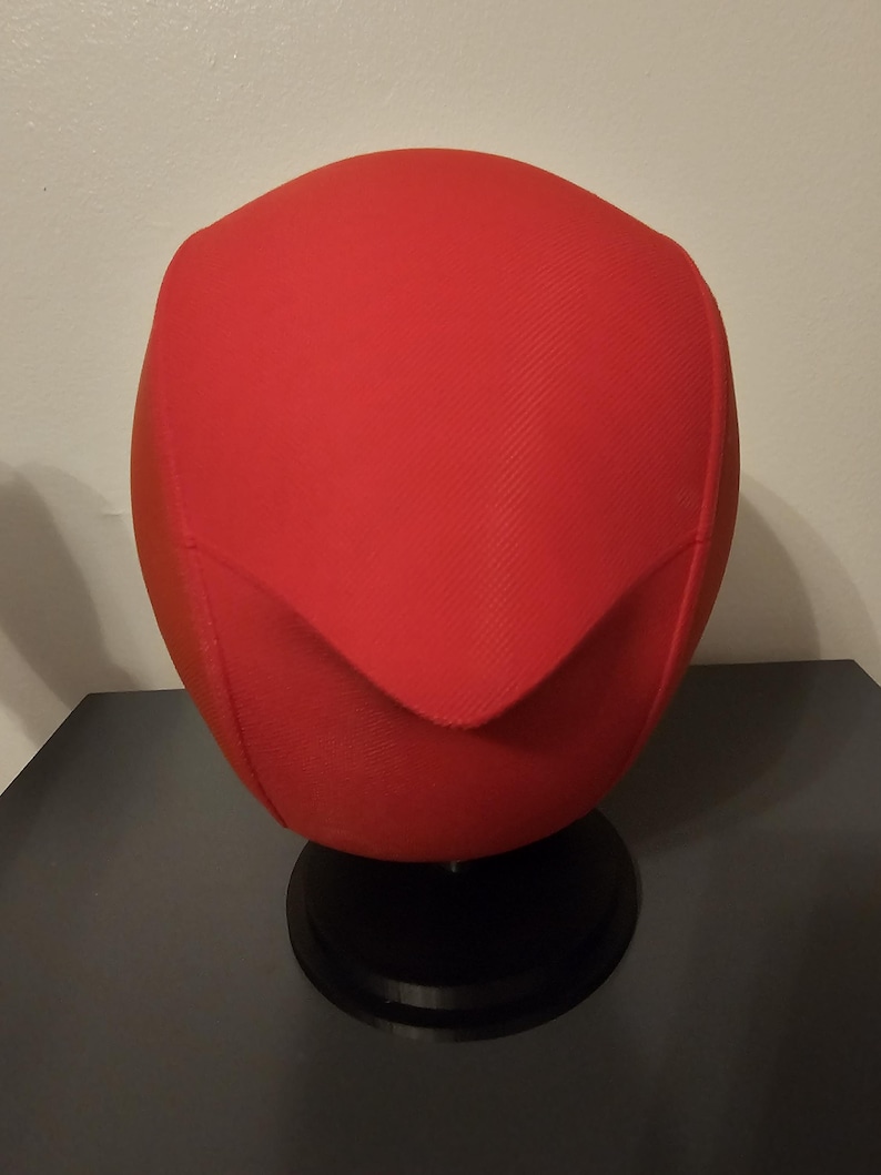 May include: A bright red, rounded object with a textured surface, possibly fabric, sits atop a black, circular base. The object has a distinct, folded-over design at the top, creating a unique shape. The base is on a dark surface.