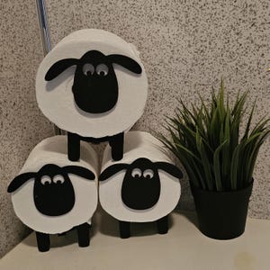 Könnte beinhalten: Drei weiße Toilettenpapierrollen mit schwarzen Schafgesichtern und Beinen, die auf einer weißen Oberfläche stehen. Rechts neben den Toilettenpapierrollen steht eine kleine grüne Topfpflanze.