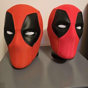 May include: Two red and black Deadpool masks. The masks have a red fabric exterior with black eye and mouth areas. The eye areas have white mesh. The masks are displayed on a gray surface.