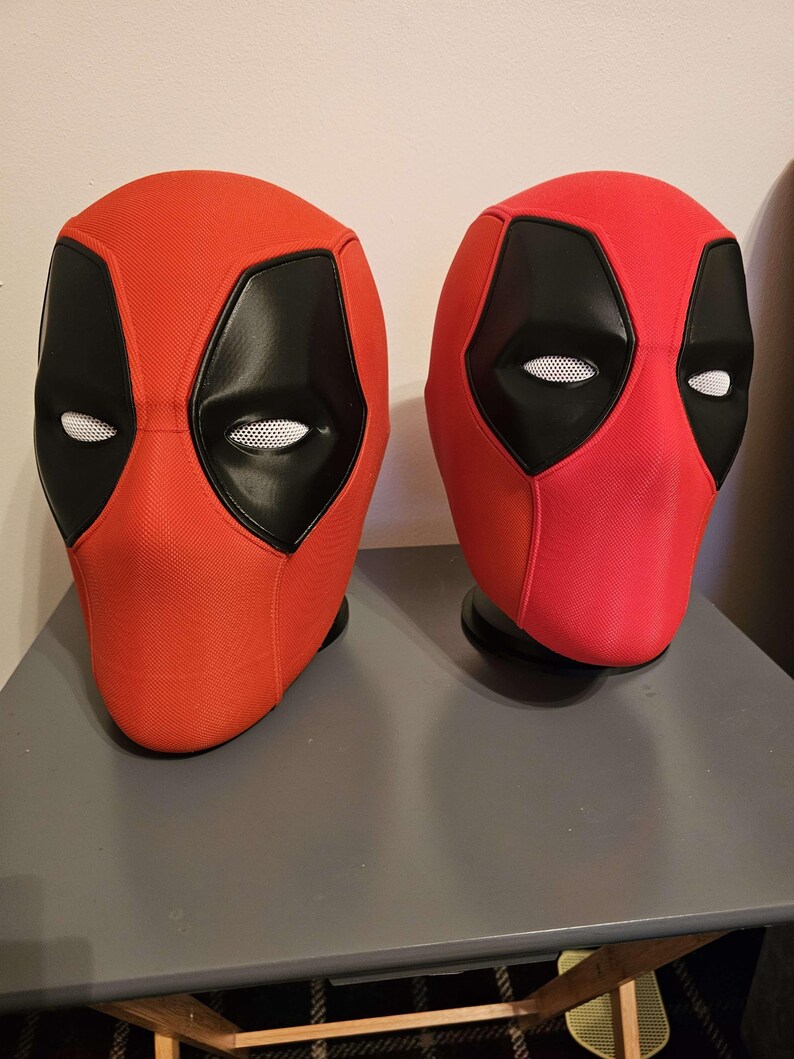May include: Two red and black Deadpool masks. The masks have a red fabric exterior with black eye and mouth areas. The eye areas have white mesh. The masks are displayed on a gray surface.