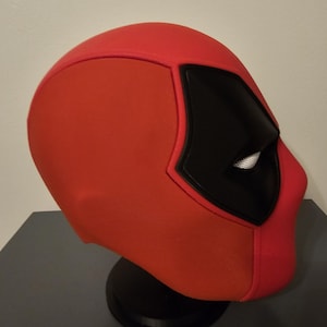 May include: A red and black Deadpool mask, a superhero costume accessory. The mask features a distinctive design with a red base, black eye coverings, and white eye details. It is displayed on a black stand.