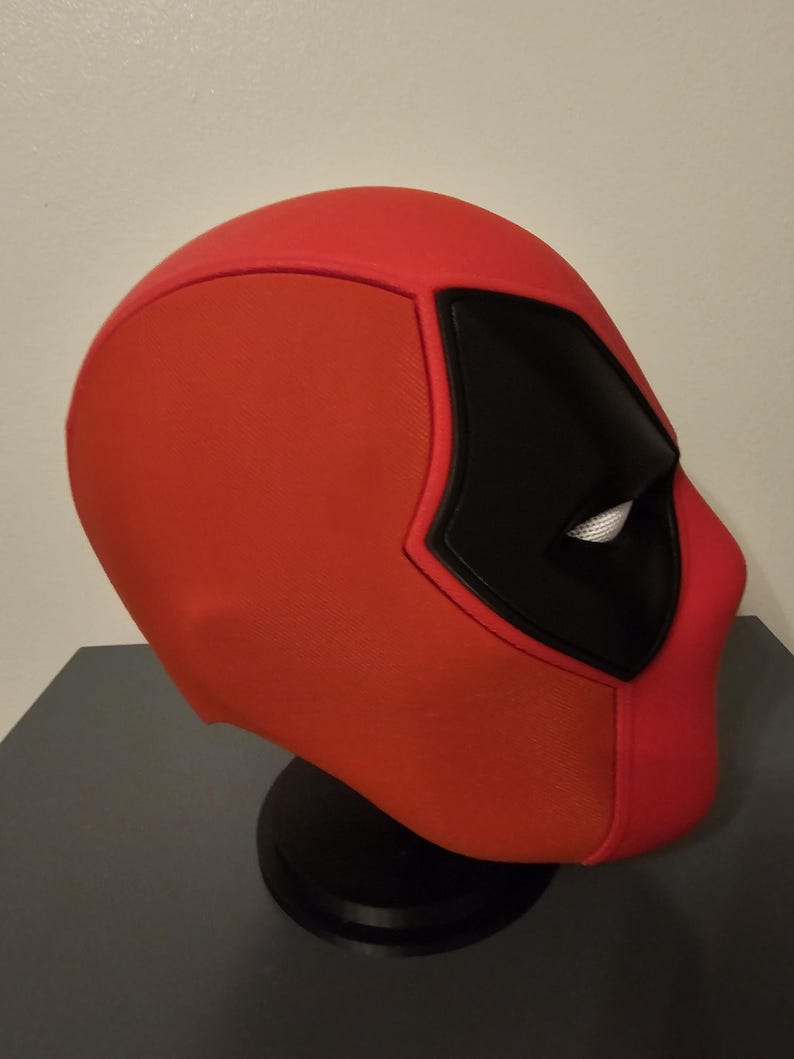 May include: A red and black Deadpool mask, a superhero costume accessory. The mask features a distinctive design with a red base, black eye coverings, and white eye details. It is displayed on a black stand.