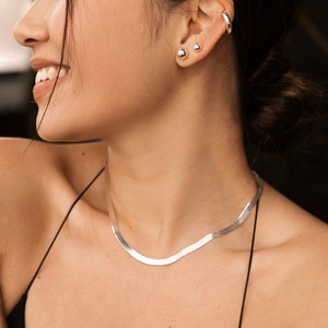 May include: A silver chain necklace with a delicate, flat, snake chain design. The necklace is worn by a person with dark hair and a silver hoop earring in one ear.