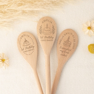 Könnte beinhalten: Drei Holzlöffel mit personalisierten Gravuren zum ersten Geburtstag. Die Löffel zeigen einen Kuchen mit einer Kerze, den Namen Amelia und den Text "1. Geburtstag Kuchen-Smash". Ein Löffel enthält auch das Datum "01.08.2024".