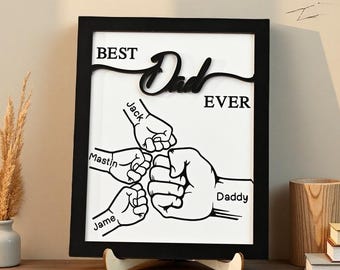 May include: A framed black and white artwork with the words "BEST Dad EVER" and the names "Jack", "Mastin", "Jame", and "Daddy" with illustrations of fists bumping. The artwork is displayed on a wooden surface.