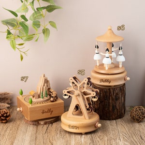 May include: Three wooden music boxes with different designs. The first music box is a mountain scene with a train, the second is a Ferris wheel, and the third is a carousel with ballerinas. Each music box has a personalized name on the base: Sally, Edith, and Debby.