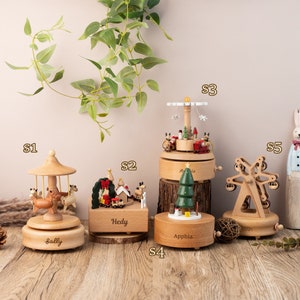 May include: Five wooden music boxes with different designs. The music boxes are decorated with Christmas themes, including a carousel, a Christmas village, a Christmas tree, a Ferris wheel, and a reindeer. The music boxes are labeled with the names Sally, Hedy, Apphia, and Erin.