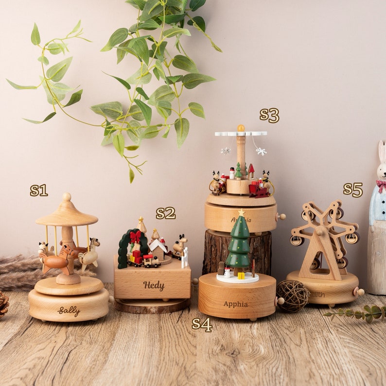 May include: Five wooden music boxes with different designs. The music boxes are decorated with Christmas themes, including a carousel, a Christmas village, a Christmas tree, a Ferris wheel, and a reindeer. The music boxes are labeled with the names Sally, Hedy, Apphia, and Erin.