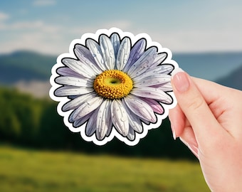 May include: A daisy sticker with white petals, a yellow center, and a black outline. The sticker has a watercolor effect and is held by a hand against a blurred background of green and blue.