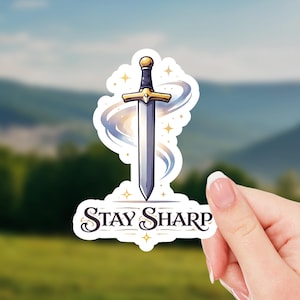 May include: A sticker featuring a stylized sword with a gold hilt and a silver blade, surrounded by a swirling blue and white design. The text "Stay Sharp" is below the sword. The sticker is held by a hand.