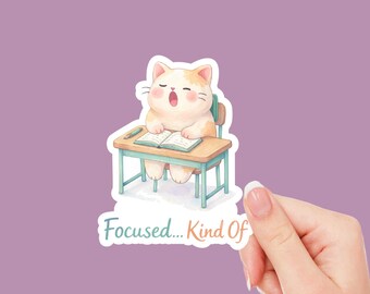 May include: A sticker featuring a cartoon cat yawning while sitting at a school desk with an open book and a pencil. The cat is white and orange. The text on the sticker reads "Focused... Kind Of".