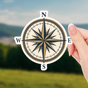 May include: A round sticker featuring a compass rose design. The compass rose has a cream-colored background with navy blue and gold accents. The cardinal directions, N, W, E, and S, are clearly marked. The sticker is held by a hand against a blurred outdoor background.