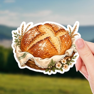 May include: A die-cut sticker featuring a round, golden-brown loaf of crusty bread with a cross-shaped score on top. The bread rests in a woven basket lined with a cream-colored cloth featuring green stripes, accented by wheat stalks and small white flowers.