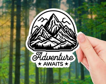 May include: A white sticker with a black mountain range design, a crescent moon, and the words "Adventure Awaits" in a decorative font. The sticker is held against a blurred forest background.