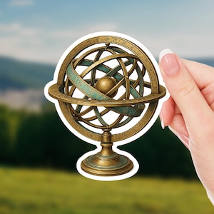 May include: A sticker featuring a vintage armillary sphere with gold and teal accents. The sphere has intricate rings and a central globe, mounted on a pedestal. The sticker is held against a blurred outdoor background.