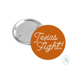 May include: A round button with a metallic silver back and a burnt orange front. The front features the words "Texas Fight!" in a white, rope-style font. A small, white logo is in the lower right corner.