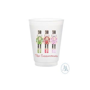May include: White plastic cup featuring three colorful nutcracker illustrations. The nutcrackers are dressed in green, red, and pink uniforms. The text "The Cussimanios" is printed in red script below the illustrations.