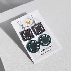 May include: Dangle earrings featuring a black square charm with the pink text KISS ALL THE TIME and a black circular charm with the blue text DISCO OCCASIONALLY. The earrings are displayed on a white card with a yellow duck logo and text.