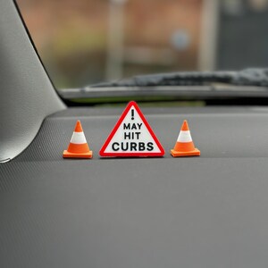 May include: A black dashboard featuring three miniature traffic cones and a warning sign. The sign is a red triangle with the words "MAY HIT CURBS" in white. The cones are orange and white.