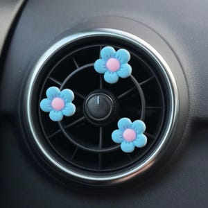 May include: Three small, five-petaled flower-shaped air vent clips attached to a circular black car air conditioning vent. Each flower features light blue petals surrounding a soft pink circular centre, creating a bright, floral accent.