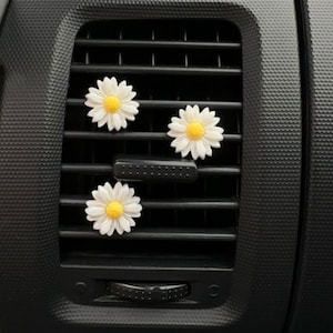 May include: Three white daisy car air vent decorations with yellow centres are attached to a black car vent. The daisies are arranged in a triangular pattern, adding a touch of floral charm to the car's interior.