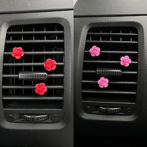 May include: Two car air vents, each with three flower-shaped air fresheners. One vent has red flowers, the other pink. The vents are black with horizontal slats, a common car interior feature.