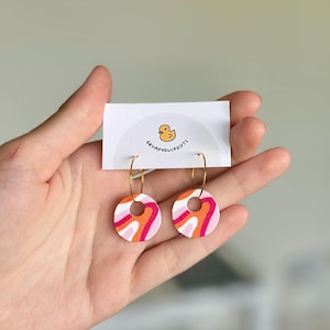 May include: A pair of hoop earrings featuring circular charms with a wavy pattern in shades of orange, pink, and white. The earrings are attached to a white display card printed with a yellow duck icon and the text GRUMPYDUCKGIFTS.