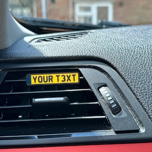 May include: A personalised car air vent tag, featuring the text "YOUR T3XT" in black on a yellow background. The rectangular tag has a blue border and is designed to fit into a car's air vent. The car's interior is black and red.