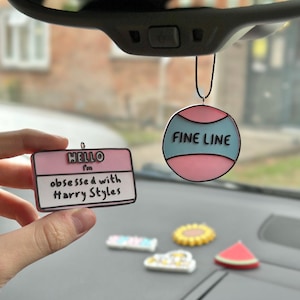 May include: Two hanging car ornaments. One is a rectangular pink and white name tag with the text "HELLO I'm obsessed with Harry Styles". The other is a circular ornament with pink and blue horizontal stripes and the text "FINE LINE".
