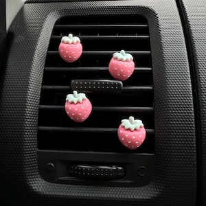 May include: Four pink strawberry car air fresheners with green tops are attached to a black car vent. The strawberries have white seed details and are evenly spaced across the vent.