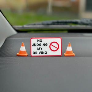 May include: A white rectangular sign with the text "NO JUDGING MY DRIVING" and a red prohibition symbol. Two small orange and white traffic cones are either side of the sign, on a grey surface.