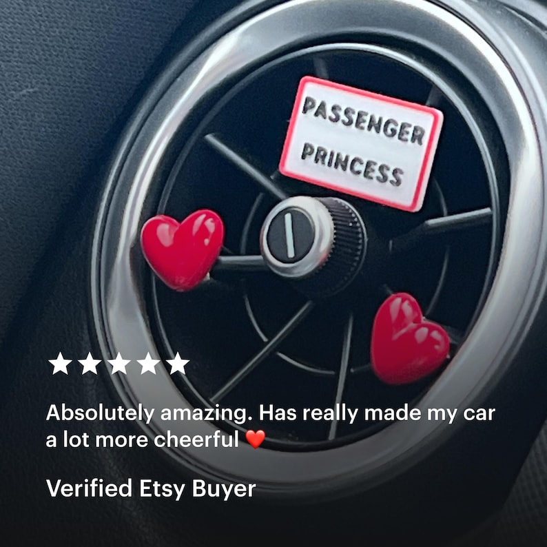 May include: Car air vent accessories featuring a red heart and a rectangular sign that reads "PASSENGER PRINCESS" in black text. The accessories are placed on a car's air vent, with a black and silver design.