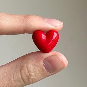May include: A small, glossy red heart-shaped object. The heart is held between two fingers, showcasing its smooth, reflective surface. The image is a close-up, highlighting the heart's vibrant color and form. The heart is a symbol of love and affection.
