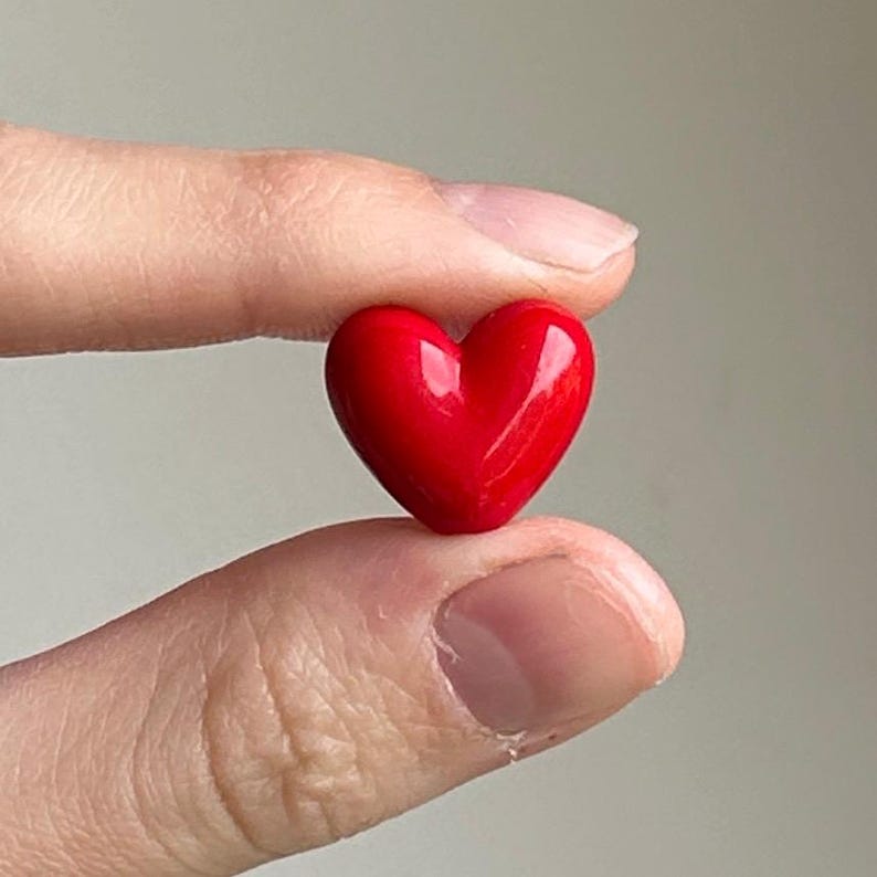 May include: A small, glossy red heart-shaped object. The heart is held between two fingers, showcasing its smooth, reflective surface. The image is a close-up, highlighting the heart's vibrant color and form. The heart is a symbol of love and affection.