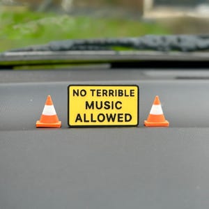 May include: A yellow sign with black text reading "NO TERRIBLE MUSIC ALLOWED" is positioned between two small orange and white traffic cones. The sign and cones are on a grey surface, with a blurred green background.