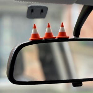 May include: Three small orange and white traffic cones sit on a car's rearview mirror. The cones are arranged in a row. The mirror has a black frame. The background is out of focus.