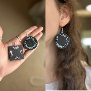 May include: Black disc earrings with the text "OCCASIONALLY. DISCO" in a light blue font. A square earring with the text "KISS ALL THE TIME" is also visible. The earrings are hanging from silver hooks.