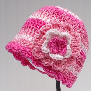 May include: A handmade crochet hat in shades of pink and white, featuring a scalloped brim and a large, three-dimensional flower. The hat is designed with a striped pattern and is likely for a child or baby.