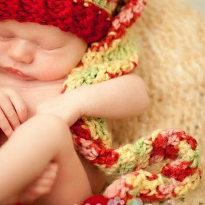 Può includere: Un neonato con un cappello e una sciarpa all'uncinetto fatti a mano. Il cappello è principalmente rosso con accenti di giallo, verde e rosa. La sciarpa è avvolta attorno al bambino, creando un aspetto accogliente e caldo. L'immagine è morbida e incentrata sul bambino.