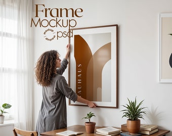 May include: A woman hangs a framed print with an abstract design in brown and white colors. The print has the word "BAUHAUS" written vertically on the side. The frame is made of wood and has a light brown finish.