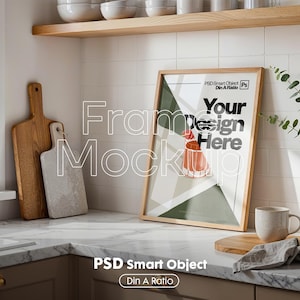 Könnte beinhalten: Ein gerahmter Kunst-Mockup mit dem Text "Your Design Here" in einer Küchenumgebung. Der Rahmen ist hellbraun, und das Kunstwerk zeigt geometrische Formen und eine rote Illustration. Ein Holzschneidebrett und eine Kaffeetasse sind ebenfalls zu sehen.