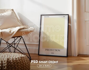 May include: A framed poster with a black border, featuring a pale yellow background and the word "PROTECTION" in black text. The poster is propped against a wall next to a chair with a beige cushion and a woven ottoman.