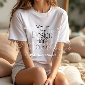 May include: A white t-shirt with the text "Your Design Here" and "PSD Smart Object" printed on it. The shirt is being worn by a person sitting on a bed.