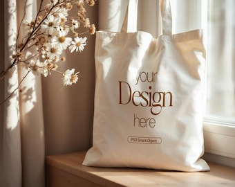 May include: A white canvas tote bag with the text "your Design here" and "PSD Smart Object" printed in brown. The bag is sitting on a wooden surface near a window.