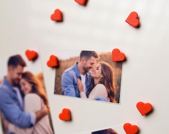 May include: Photos of a couple embracing, held in place by small, textured red heart magnets. The photos are on a white surface, with several heart magnets scattered around the images, creating a romantic theme.