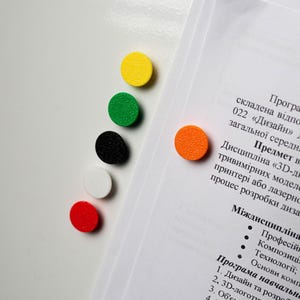 Może przedstawiać: Kolekcja kolorowych, okrągłych magnesów w kolorze żółtym, zielonym, czarnym, białym i czerwonym jest ułożona na białej powierzchni. Pomarańczowy magnes jest przymocowany do dokumentu z tekstem. Magnesy są prawdopodobnie używane do organizowania lub oznaczania.