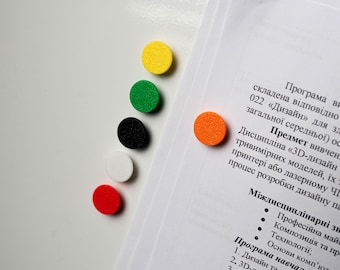 May include: A collection of colorful, round magnets in yellow, green, black, white, and red are arranged on a white surface. An orange magnet is attached to a document with text. The magnets are likely used for organizing or marking.